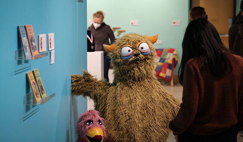 A small fuchsia duck-like puppet and a large brown monster puppet are positioned against a teal and blue background in a museum gallery