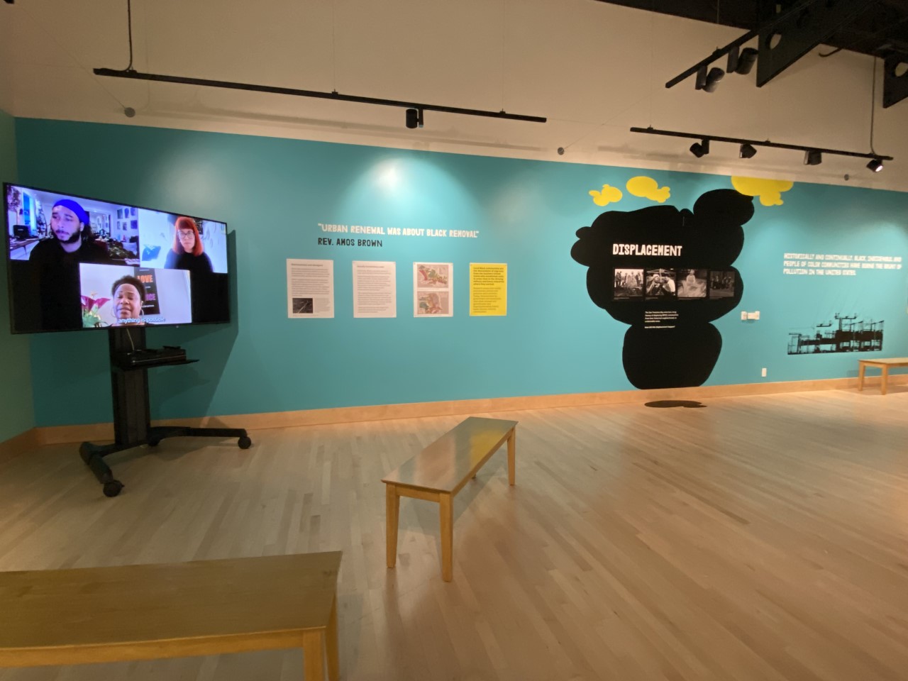 A TV is at the corner of a blue-green wall playing a video of two students interviewing a community member. To the right of the TV are four posters under a quote that reads urban renewal was about black removal, reverend Amos Brown. Next to the posters is a large black cloud like graphic with bright yellow accents, and Displacement in white vinyl letters. Four posters underneath this title are historical images of African Americans during the World War two era from the SF State Labor Archives.