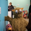 A small fuchsia duck-like puppet and a large brown monster puppet are positioned against a teal and blue background in a museum gallery