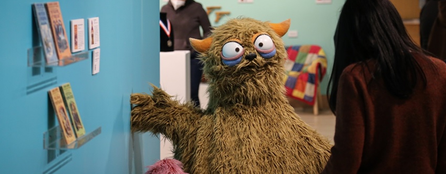 A small fuchsia duck-like puppet and a large brown monster puppet are positioned against a teal and blue background in a museum gallery