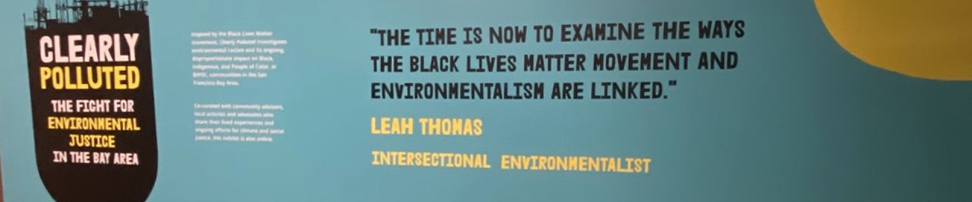 On a blue green wall is an oil barrel with the words Clearly Polluted The Fight for Environmental Justice in the Bay Area, and a quote in chunky black letters that reads the time is now to examine the ways the black lives matter movement and environmentalism are linked by Leah thomas. At the top right of the wall are bright yellow toxic clouds.