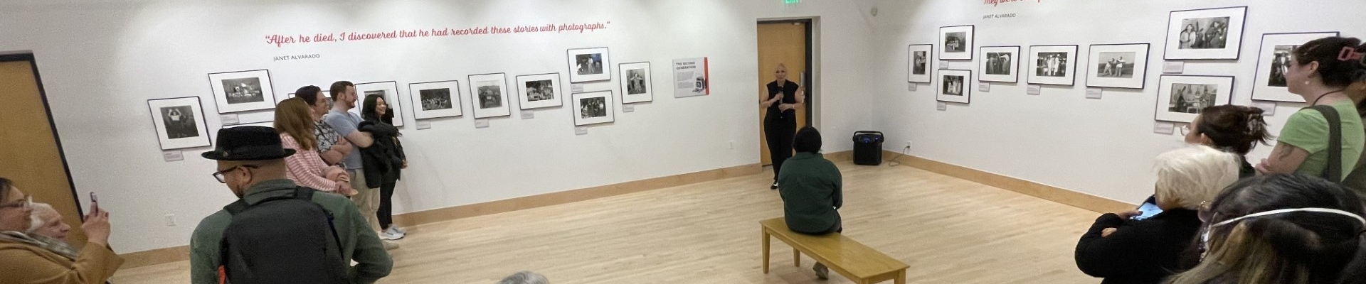 a group of people in a gallery listen to Janet Alvarado speaking during an opening reception of her father's photography