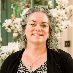 A white woman with curly salt and pepper shoulder length hair poses in front of a flower arrangement,smiling, and wearing a black cardigan over a v-neck black/white flower pattern top
