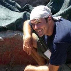 Michael smiling, kneeling down next to an excavation site, wearing a white bandana
