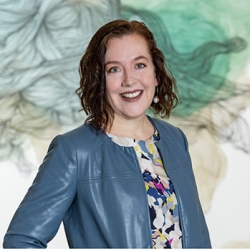 A smiling white woman with red-brown hair wearing a blue leather coat stands in front of a blue and teal wall painting 