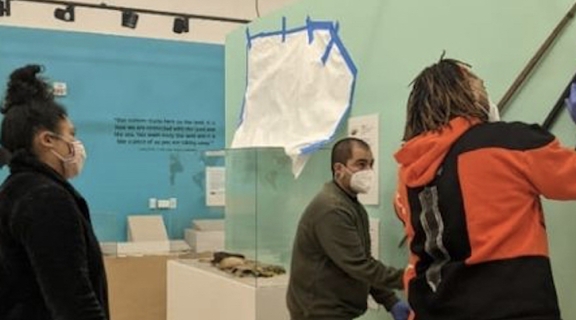 1 Latina female, 1 Latino male and 1 African American male student work to remove wood bows from a gallery wall