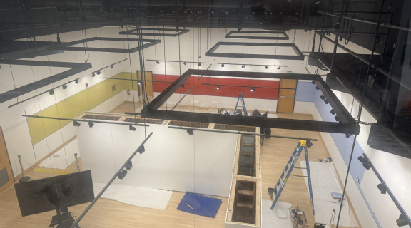 Overhead view of the Global Museum during exhibition installation, showing partially constructed gallery walls, display cases, ladders, painting supplies, and protective floor coverings