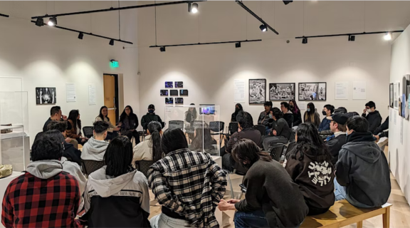 Asian American studies professors hold a discussion with students in a museum gallery with warm white walls and a selection of photographs displayed on a wall