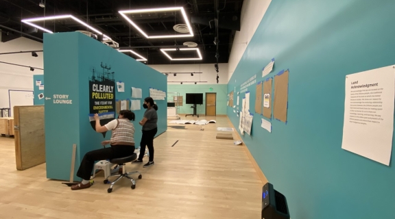 Two students apply black vinyl signace of an oil barrel onto a blue-green wall